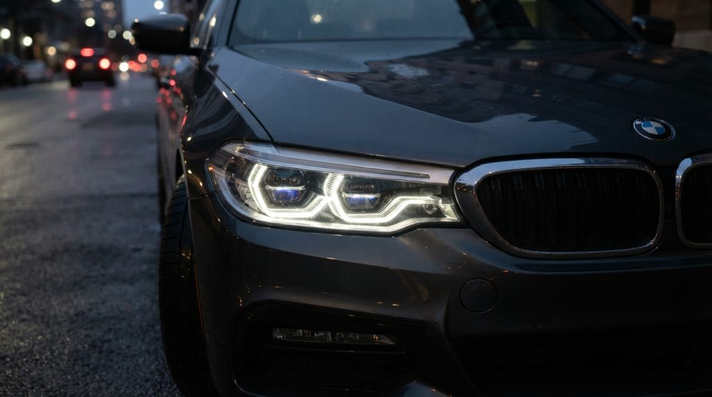 Na tym obrazie widać realistyczne zbliżenie na przód nowoczesnego samochodu osobowego (marki BMW) zaparkowanego na miejskiej ulicy o zmierzchu lub w nocy